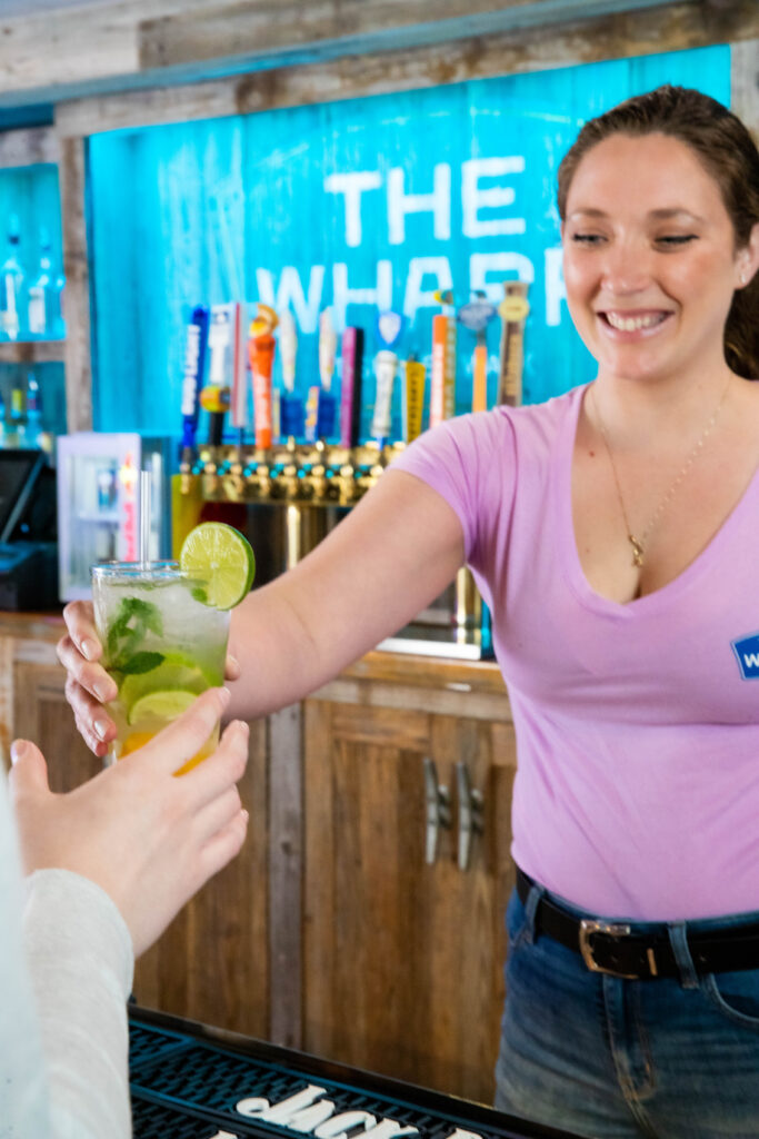 Smiling woman bartender hands a cocktail with sliced lime to a guest at The Wharf